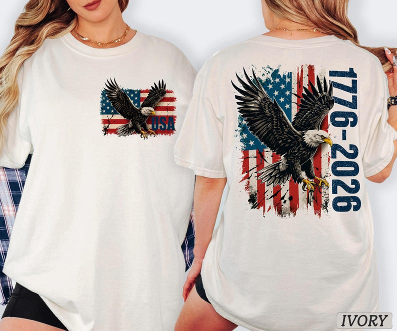 Pode incluir: Uma t-shirt cor de marfim com o gr&aacute;fico de uma &aacute;guia-de-cabe&ccedil;a-branca a voar sobre uma bandeira americana desgastada. O peito apresenta uma pequena &aacute;guia e a bandeira com o texto USA. As costas mostram uma grande &aacute;guia, a bandeira e o texto 1776-2026 em azul.