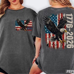 Pode incluir: Uma t-shirt cinzento-carv&atilde;o com um gr&aacute;fico de uma &aacute;guia-de-cabe&ccedil;a-branca a voar sobre uma bandeira americana desgastada. O peito exibe a &aacute;guia e o texto USA. As costas apresentam uma &aacute;guia maior, a bandeira e o texto 1776-2026.