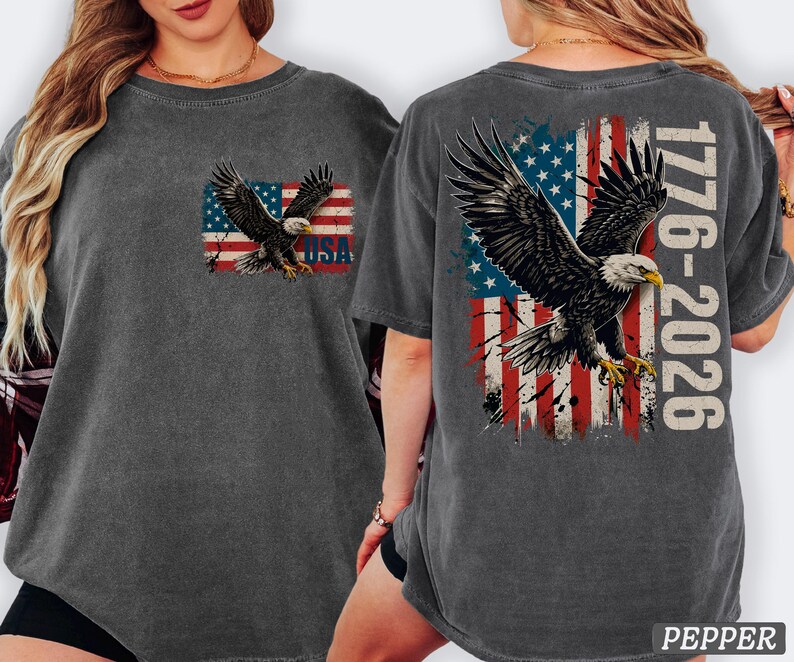 Pode incluir: Uma t-shirt cinzento-carv&atilde;o com um gr&aacute;fico de uma &aacute;guia-de-cabe&ccedil;a-branca a voar sobre uma bandeira americana desgastada. O peito exibe a &aacute;guia e o texto USA. As costas apresentam uma &aacute;guia maior, a bandeira e o texto 1776-2026.