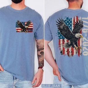 Pode incluir: Uma t-shirt azul com um gr&aacute;fico no peito esquerdo de uma &aacute;guia-de-cabe&ccedil;a-branca voando sobre uma bandeira americana desgastada com o texto USA. As costas exibem uma vers&atilde;o maior do mesmo gr&aacute;fico com o texto 1776-2026.