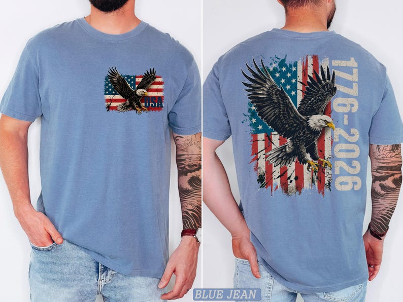 Pode incluir: Uma t-shirt azul com um gr&aacute;fico no peito esquerdo de uma &aacute;guia-de-cabe&ccedil;a-branca voando sobre uma bandeira americana desgastada com o texto USA. As costas exibem uma vers&atilde;o maior do mesmo gr&aacute;fico com o texto 1776-2026.