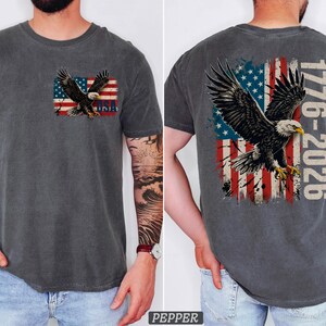 Pode incluir: Uma t-shirt cinzento-carv&atilde;o de manga curta com o gr&aacute;fico de uma &aacute;guia-de-cabe&ccedil;a-branca a voar sobre uma bandeira americana desgastada. O peito exibe uma pequena &aacute;guia e o texto USA, as costas uma &aacute;guia grande e o texto 1776-2026.