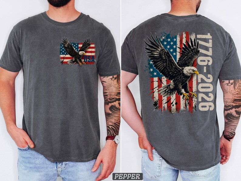 Pode incluir: Uma t-shirt cinzento-carv&atilde;o de manga curta com o gr&aacute;fico de uma &aacute;guia-de-cabe&ccedil;a-branca a voar sobre uma bandeira americana desgastada. O peito exibe uma pequena &aacute;guia e o texto USA, as costas uma &aacute;guia grande e o texto 1776-2026.