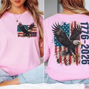Pode incluir: Uma t-shirt rosa claro com um gr&aacute;fico de uma &aacute;guia-de-cabe&ccedil;a-branca voando sobre uma bandeira americana desgastada. O peito exibe a &aacute;guia e a bandeira com o texto USA. As costas apresentam uma vers&atilde;o maior com a &aacute;guia, a bandeira e 1776-2026.