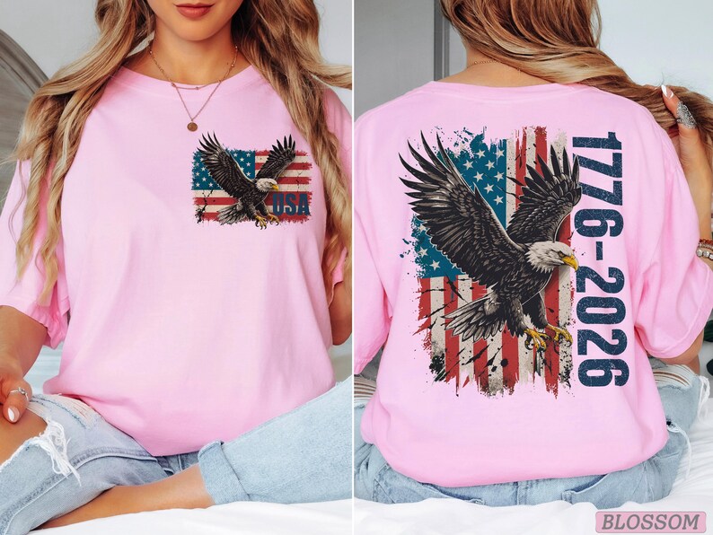 Pode incluir: Uma t-shirt rosa claro com um gr&aacute;fico de uma &aacute;guia-de-cabe&ccedil;a-branca voando sobre uma bandeira americana desgastada. O peito exibe a &aacute;guia e a bandeira com o texto USA. As costas apresentam uma vers&atilde;o maior com a &aacute;guia, a bandeira e 1776-2026.