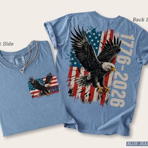 Pode incluir: Uma t-shirt azul jeans mostrada de frente e de costas. A frente apresenta um pequeno gr&aacute;fico de uma &aacute;guia sobre uma bandeira americana e o texto USA. As costas exibem uma &aacute;guia grande, uma bandeira americana desgastada e o texto 1776-2026.