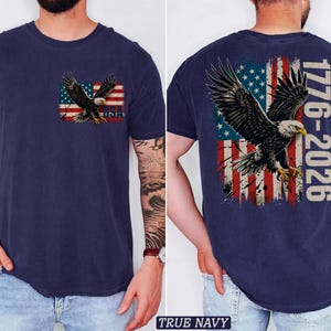 Pode incluir: Uma t-shirt azul-marinho com um gr&aacute;fico no peito esquerdo de uma &aacute;guia-de-cabe&ccedil;a-branca sobre uma bandeira americana desgastada com o texto USA. As costas exibem uma vers&atilde;o maior do mesmo motivo com o texto 1776-2026.