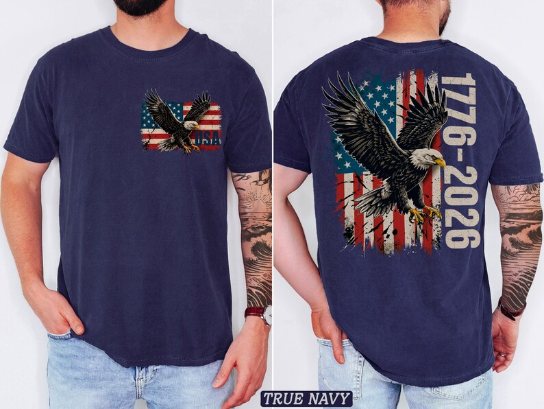 Pode incluir: Uma t-shirt azul-marinho com um gr&aacute;fico no peito esquerdo de uma &aacute;guia-de-cabe&ccedil;a-branca sobre uma bandeira americana desgastada com o texto USA. As costas exibem uma vers&atilde;o maior do mesmo motivo com o texto 1776-2026.