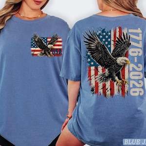 Pode incluir: Uma t-shirt azul com um gr&aacute;fico no peito esquerdo de uma &aacute;guia-de-cabe&ccedil;a-branca sobre uma bandeira americana desgastada com o texto USA. As costas exibem uma grande &aacute;guia em voo sobre uma bandeira americana com o texto 1776-2026.
