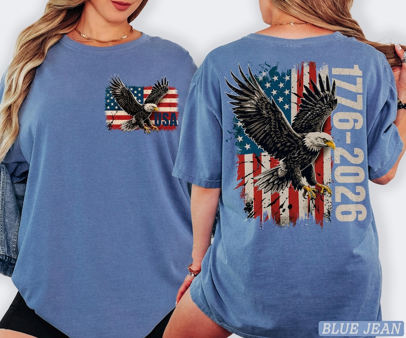 Pode incluir: Uma t-shirt azul com um gr&aacute;fico no peito esquerdo de uma &aacute;guia-de-cabe&ccedil;a-branca sobre uma bandeira americana desgastada com o texto USA. As costas exibem uma grande &aacute;guia em voo sobre uma bandeira americana com o texto 1776-2026.