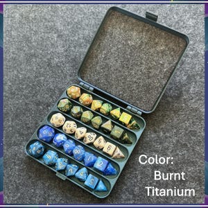 May include: A dark gray rectangular storage case containing a collection of multi-sided gaming dice arranged in rows. The dice feature various colors including blue, cream, and green tones. The text Color: Burnt Titanium is visible.