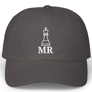 Puede incluir: Gorra de béisbol gris oscuro con un rey de ajedrez blanco y las letras "MR" debajo. La gorra tiene una visera curva y un diseño clásico de seis paneles. Un accesorio elegante para los entusiastas del ajedrez.