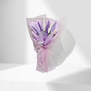 May include: A bouquet of lavender-coloured flowers, wrapped in light purple paper and tied with a pink ribbon. The bouquet features a mix of purple and white flowers, creating a soft and delicate appearance. The background is plain white.
