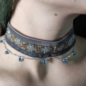Può includere: Una collana girocollo in velluto grigio con perline dorate e blu. La collana ha una delicata catena d'argento con pendenti blu a forma di goccia. Il girocollo è indossato intorno al collo.