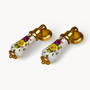 May include: Two decorative cabinet pulls. Each pull has a white porcelain handle decorated with yellow and purple floral patterns. The handles are accented with gold-coloured metal hardware, including a decorative backplate and mounting hardware.