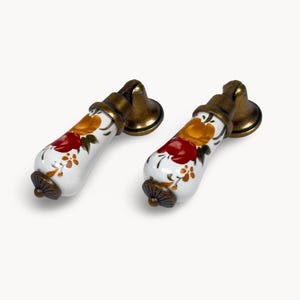 May include: A pair of cabinet pulls. Each pull has a white ceramic handle decorated with red and yellow floral patterns. The metal parts are brass-coloured. These decorative pulls would add a vintage touch to furniture.
