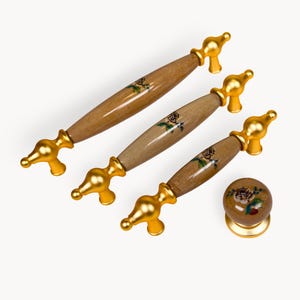 May include: A set of cabinet hardware featuring three handles and a knob. The handles have a light brown wooden centre with a painted rose design and gold-coloured metal accents. The knob has a similar design.