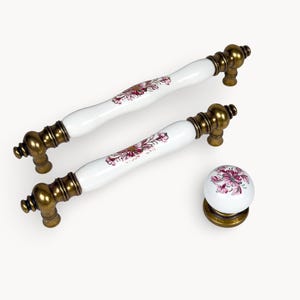 May include: Two white and gold cabinet pulls and a matching knob. The pulls have a white ceramic centre with a floral design in shades of pink and burgundy. The hardware is a warm gold colour.