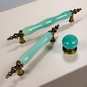 May include: Two aqua-coloured cabinet handles and a matching knob with antique brass hardware. The handles have a curved, ceramic design. The knob is a round, ceramic sphere. The hardware has an ornate design.