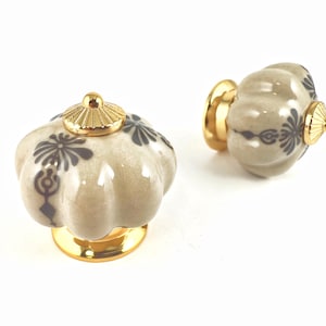 May include: Two ceramic cabinet knobs with a pumpkin shape. The knobs are light gray with dark gray floral patterns and gold-colored accents. The knobs have a gold base and a gold cap on top.