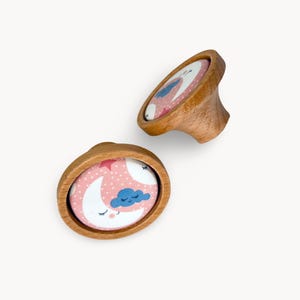 Peut inclure: Deux boutons de meuble en bois avec un design fantaisiste. Chaque bouton présente un fond rose avec un croissant de lune, un nuage et des étoiles. Les boutons sont en bois de couleur claire.