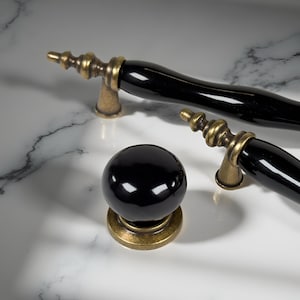 May include: Close-up of black and brass cabinet hardware. The set includes two curved handles and a round knob. The hardware has a glossy black finish with brass accents. The items are displayed on a white marble surface.