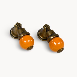 Può includere: Due pomelli per mobili dall'estetica vintage. Ogni pomello presenta un pomolo sferico in ceramica arancione brillante, montato su una base e ferramenta in metallo color bronzo. La ferramenta ha un aspetto ornato e invecchiato.