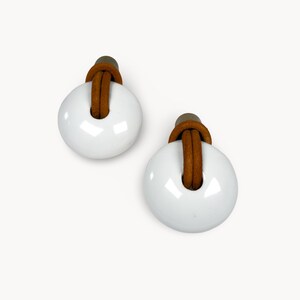 May include: Two white, circular earrings with a glossy finish. Each earring features a brown leather strap looped through a central hole and attached to a small, dark brown post. The earrings are set against a white background.