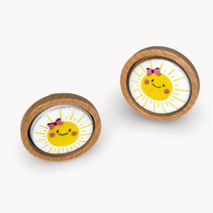 May include: A pair of wooden stud earrings. Each earring features a smiling yellow sun with a pink bow, set against a white background. The design is framed by a light brown wooden border. A bright and cheerful accessory.