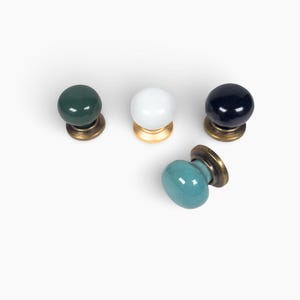 May include: Four decorative cabinet knobs in various colors: dark green, white, navy blue, and teal. Each knob has a rounded ceramic top and a brass base. The knobs are arranged on a white background.