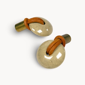 May include: A pair of cufflinks with a light beige, oval stone design, brown leather knot detail, and dark brass cylindrical post. The cufflinks are set against a white background. The stone design is approximately 2 cm in diameter.