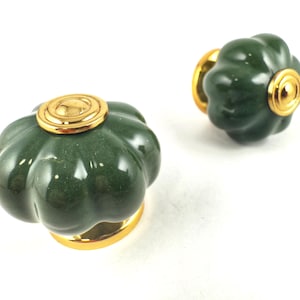 May include: Two dark green cabinet knobs with a flower-like design. Each knob has a gold-coloured base and a gold-coloured circular accent on the top. The knobs are set against a white background.