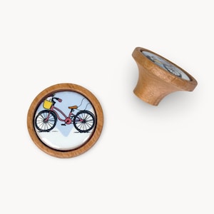 Puede incluir: Dos pomos de armario de madera con diseño de bicicleta. Los pomos tienen una base redonda de madera clara con una ilustración de bicicleta en el centro. La bicicleta es rosa con ruedas negras y una cesta amarilla. Un pomo está en posición vertical, el otro en ángulo.