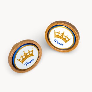 Peut inclure: Deux boutons de meuble en bois avec un centre ovale blanc orné d'une couronne dorée et du mot "Prince" en bleu. Les boutons ont un bord bleu et un bord en bois naturel.
