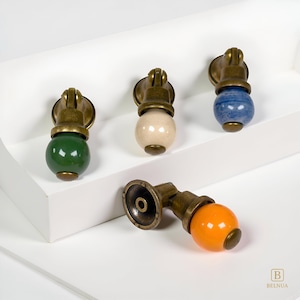 May include: A collection of cabinet pulls with brass-toned hardware and colorful ceramic spheres. The pulls feature green, cream, blue, and orange spheres. The hardware has a decorative design. The pulls are displayed on a white surface.
