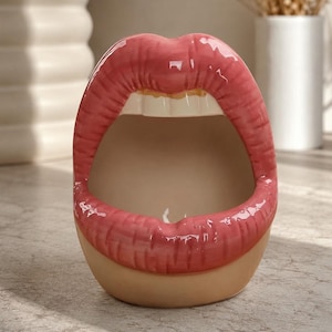 May include: A ceramic container shaped like an open mouth with glossy pink lips and visible white teeth. The interior of the mouth is a smooth, light beige colour, and the base of the object is a solid, rounded beige form, creating a functional vessel.