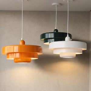 Può includere: Tre lampade a sospensione a livelli appese a cavi bianchi. Una lampada è arancione brillante, una è verde bosco scuro e una è bianca. Ogni apparecchio presenta un design circolare a strati con una finitura liscia e lucida.