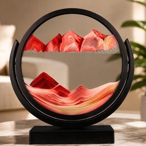 Op de afbeelding: Een cirkelvormig zwart frame bevat een dynamisch zandkunstwerk met lagen rood, roze en wit zand. Het zand vormt bergachtige pieken en dalen in het glas, met een horizontale lijn van kleine luchtbelletjes die de bovenste en onderste secties scheidt.