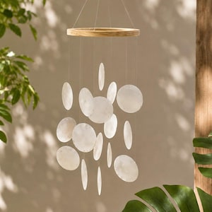 Peut inclure: Un mobile suspendu composé d'un cadre circulaire en bois clair auquel sont suspendus de multiples disques circulaires en coquillage blanc translucide par de fins filaments transparents à des longueurs variées, créant un effet en cascade.