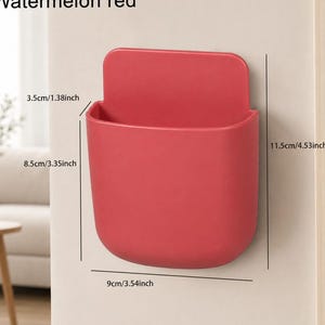 Könnte beinhalten: Eine wassermelonenrote Wandaufbewahrungstasche in abgerundeter rechteckiger Form. Die Maße sind mit 3,5 cm Tiefe, 8,5 cm Breite und 11,5 cm Höhe angegeben. Der Behälter verfügt über eine flache Rückwand zur Montage und eine offene Oberseite.