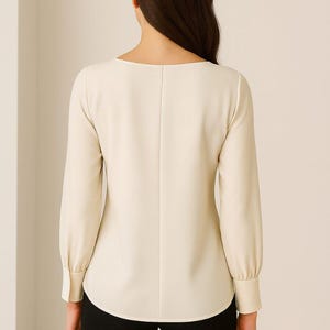 May include: Cream-colored long-sleeve blouse with a scoop neckline and buttoned cuffs. The back of the blouse features a vertical seam down the center. The bottom half of the image shows black pants.