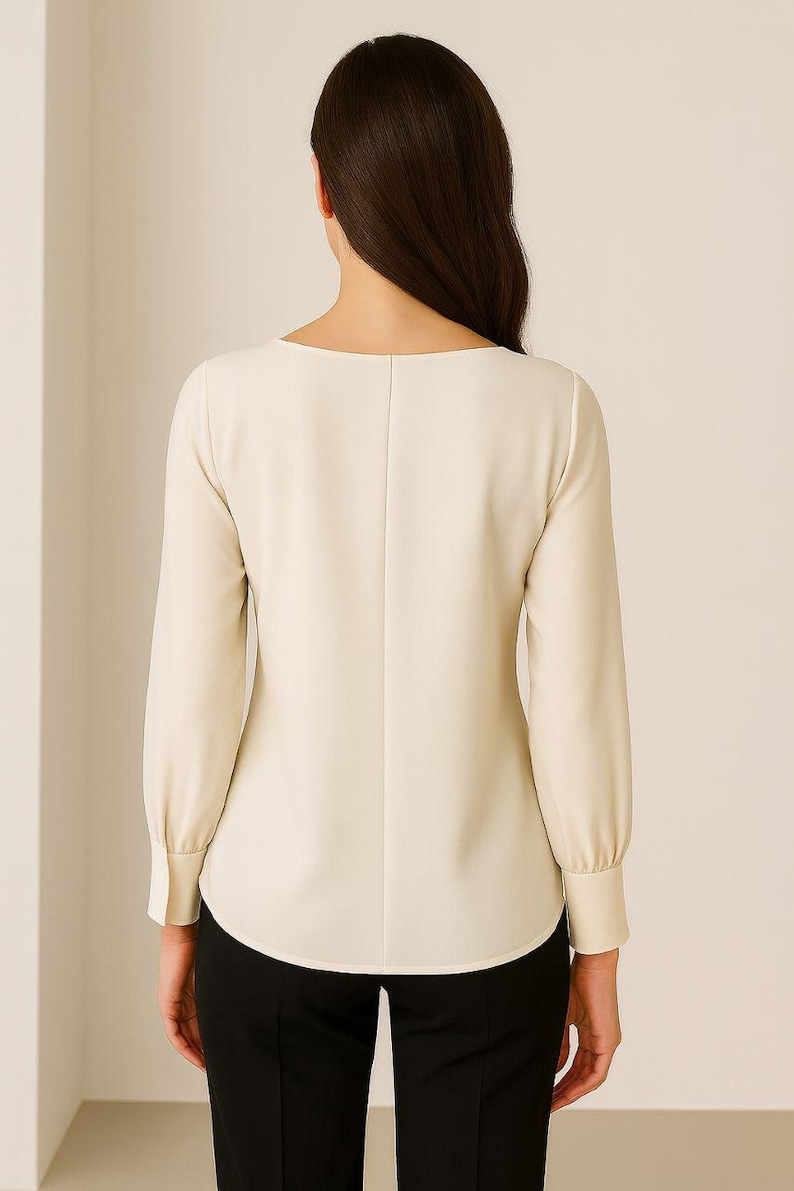 May include: Cream-colored long-sleeve blouse with a scoop neckline and buttoned cuffs. The back of the blouse features a vertical seam down the center. The bottom half of the image shows black pants.