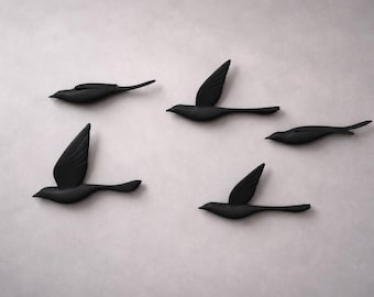 Puede incluir: Un conjunto de cinco esculturas de pared de aves en color negro mate dispuestas en formación de bandada. Cada ave se muestra en una postura de vuelo diferente, algunas con las alas extendidas hacia arriba y otras con las alas plegadas.