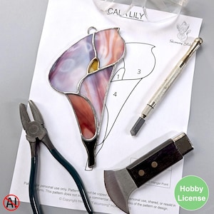 May include: A stained glass cala lily with purple, pink, and yellow hues, next to a pattern, pliers, a glass cutter, and a knife. The pattern is labeled "CALA LILY" and includes the text "Falconsong Studio". A "Hobby License" sticker is in the corner.