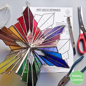 May include: A multi-colored stained glass star ornament with a silver metal frame, positioned over an Art Deco Spinner pattern sheet. The assembly includes a glass cutter, red-handled scissors, and pliers, showcasing the craft process.