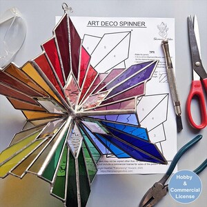 May include: A multi-colored stained glass Art Deco spinner ornament featuring red, orange, yellow, green, blue, and purple panels joined by silver solder. The piece is displayed alongside a printed pattern sheet, a glass cutter, and metal pliers.