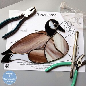 May include: A stained glass Canada goose suncatcher featuring brown, black, and white glass pieces joined by dark solder lines. The piece rests on a printed paper pattern labelled CANADA GOOSE, accompanied by metal pliers, a glass cutter, and safety glasses.