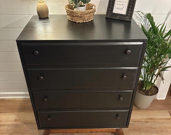 May include: A black wooden dresser with four drawers and dark knobs. The dresser has a mid-century modern design with angled wooden legs. A small lamp, a woven basket with a plant, and a framed print are on top.