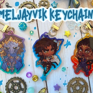 Op de afbeelding: Sleutelhangers met kleurrijke cartoonkarakterontwerpen en goudkleurige hardware. De sleutelhangers hebben een verscheidenheid aan kleuren, waaronder blauw, oranje en paars. De tekst "MELJAYVIK KEYCHAINS" wordt weergegeven in een lichtblauwe letter.