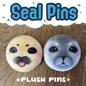 May include: Two plush seal pins. One is light yellow with a pink blush and a black nose and eyes. The other is light blue with a black nose and eyes. The text "Seal Pins" and "Plush Pins" are in the image.